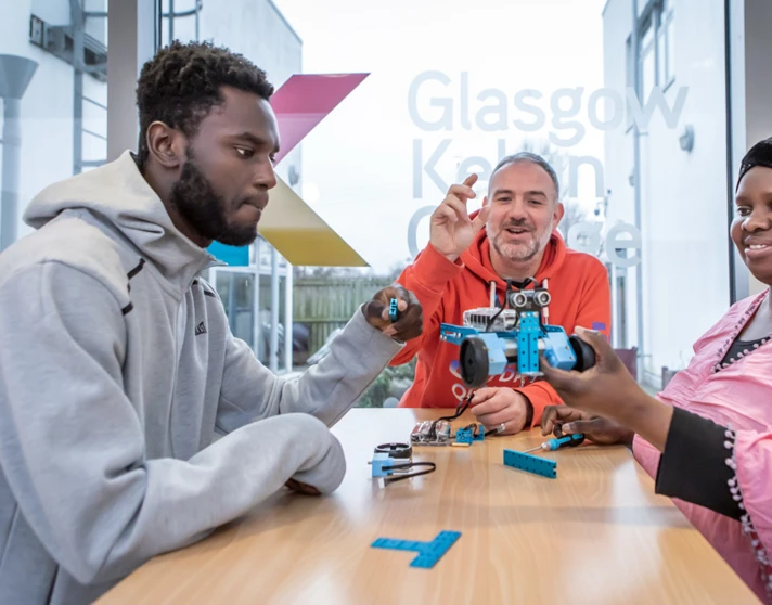 Lecturer in an orange hoodie helping two students troubleshoot and assemble a blue robotic vehicle in a classroom. Lecturer in an orange hoodie helping two students troubleshoot and assemble a blue robotic vehicle in a classroom.